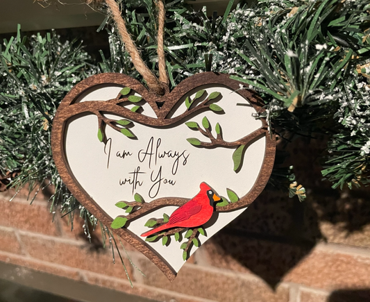 Heart shaped ornament with white background and tree branches with leaves in the shape of a heart. Leaves are painted green and a red cardinal sits on the branches. “I am always with you” is engraved on the background.
